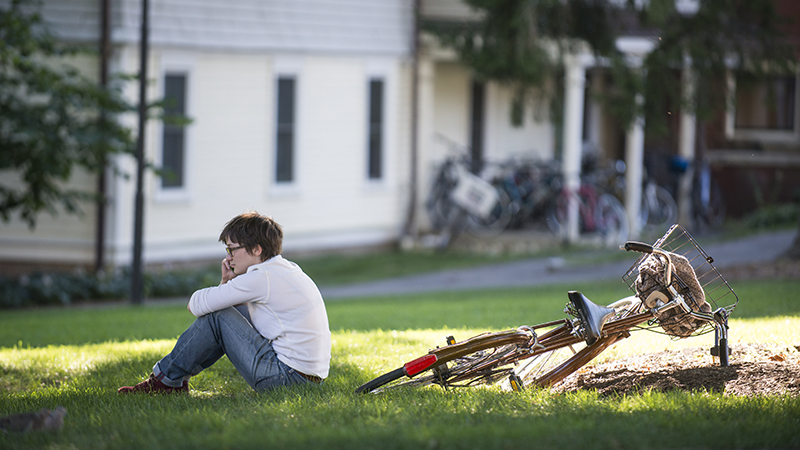 Student sitting on grass, talking on cell phone, with bicycle leaning on grass behind her