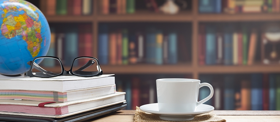 A globe centered on Africa sits on the corner of a desk. A pile of books is topped by a pair of glasses and to one side a saucer with coffee or tea. A vast bookcase covers the wall.