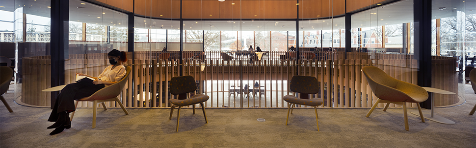 Photo of a masked woman sitting in a chair inside of the new Neilson library, reading a book. Students study in the distance. 