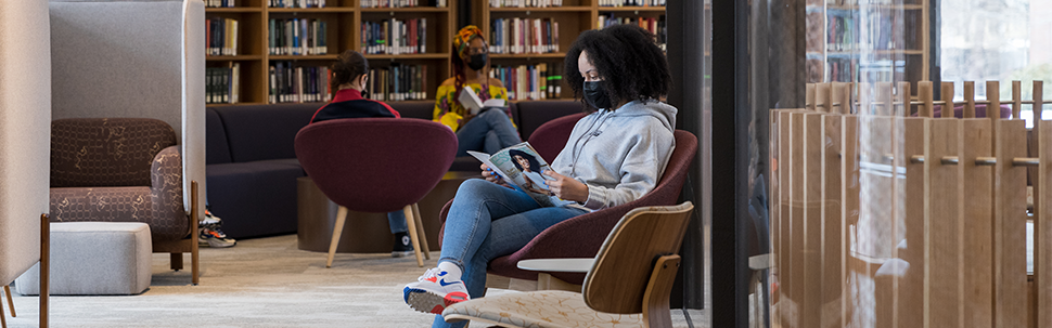 Student sitting in a chair reading a magazine, while a group of students studies at a table in the background.