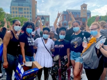 Six friends—five black and one white— in masks standing next to each other in front of a citscape. T-shirts include "Black Nerds Unite" and "Melanin Poppin"