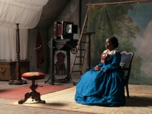 Color film still of a Black woman in 19th century clothing in a photo studio