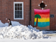 Person walking through snow drifts. Behind them is a brick building with a bright rainbow banner