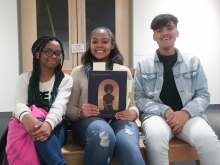 Three students of color seated and smiling at the camera. The middle person holds a copy of the exhibit book Black Refractions