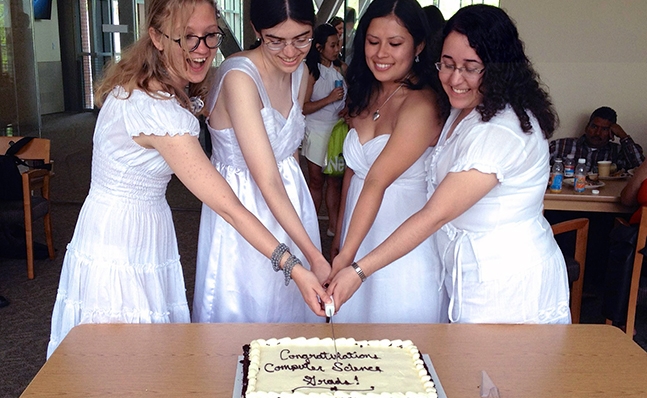 Four computer science grads in Commencement dresses cutting a cake